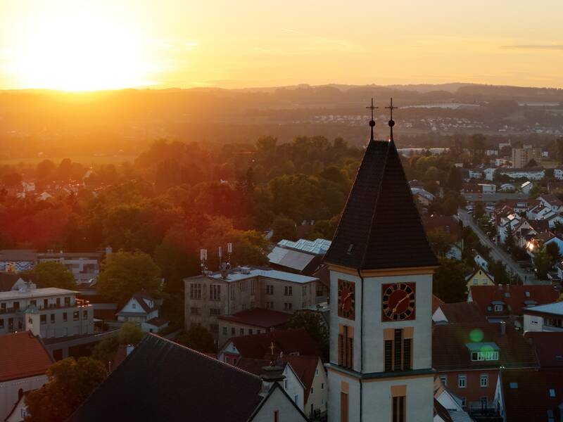 Das Bild zeigt eine Stadtlandschaft bei Sonnenuntergang mit einem Kirchturm im Vordergrund und weiten, sanft beleuchteten Hügeln im Hintergrund.