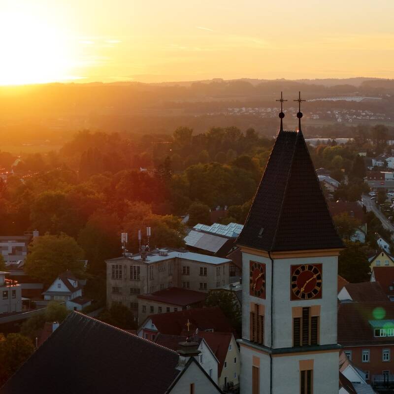 Das Bild zeigt eine Stadtlandschaft bei Sonnenuntergang mit einem Kirchturm im Vordergrund und weiten, sanft beleuchteten Hügeln im Hintergrund.