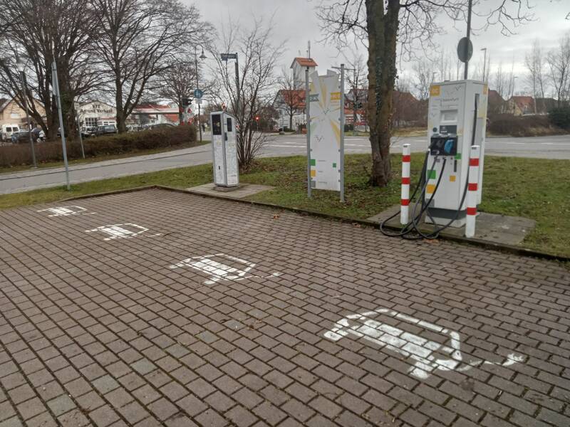Auf dem Bild sind mehrere Ladestationen für Elektroautos auf einem gepflasterten Parkplatz zu sehen, umgeben von Bäumen und einer Straße im Hintergrund.