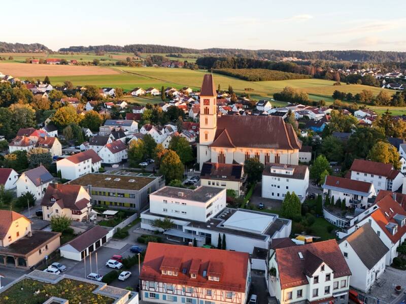 Eine Luftaufnahme zeigt eine kleine Stadtlandschaft mit einer zentralen Kirche, umgeben von Wohnhäusern, Feldern und Wäldern.
