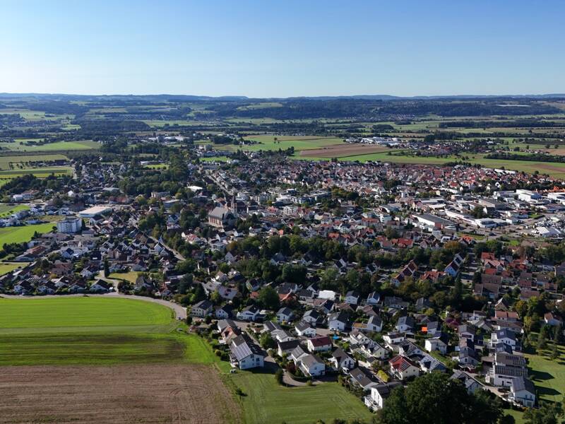 Die Aufnahme zeigt eine Luftansicht einer weitläufigen ländlichen Stadt, umgeben von grünen Feldern und Wäldern unter klarem blauem Himmel.
