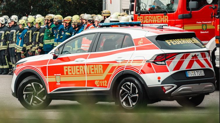 Einsatzfahrzeug der Feuerwehr fährt vor einer Gruppe Feuerwehrleute vorbei