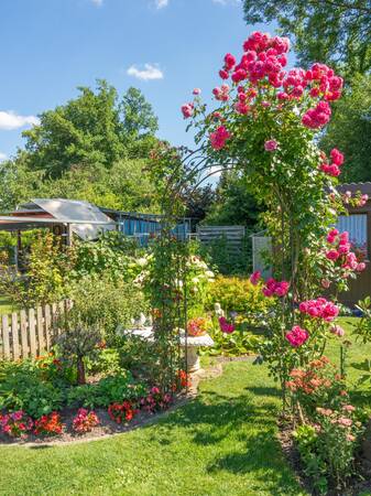 Ein üppig blühender Garten mit einem Rosenbogen und verschiedenen Blumenbeeten an einem sonnigen Tag.