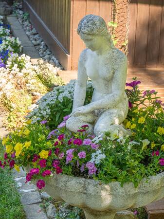 Eine Steinstatue einer knienden Frau ist von bunten Blumen umgeben und steht in einem Garten vor einer Holzwand.