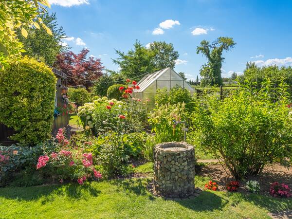 Ein lebendiger Garten mit vielen blühenden Pflanzen, einem Gewächshaus und einem kleinen Brunnen.