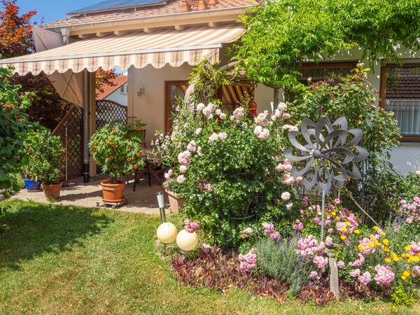 Ein sonniger Garten mit blühenden Rosen und bunten Blumen steht vor einer Terrasse mit Markise und Topfpflanzen.