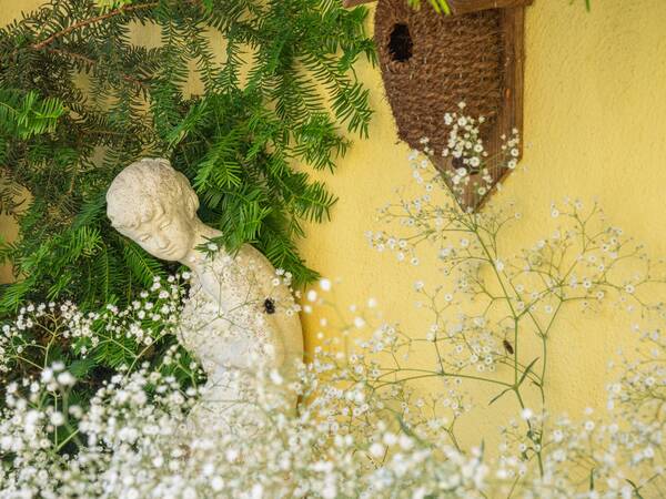 Eine steinerne Skulptur eines kopfneigenden Kindes steht umgeben von grünen Zweigen und weißen Blüten vor einer gelben Wand, an der ein kleiner, rustikaler Nistkasten hängt.