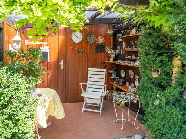 Ein gemütlicher Gartenbereich mit einer Holzwand voller Deko-Objekte, einer weißen Gartenstuhlgruppe und grüner Bepflanzung umgeben von dichten Sträuchern und einem kleinen Tisch mit gelber Tischdecke.