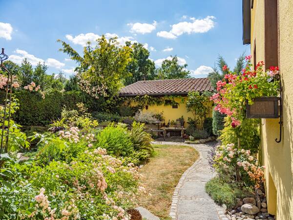 Ein gemütlicher Garten mit einem geschwungenen Steinweg, umgeben von blühenden Pflanzen, führt zu einer Sitzbank vor einem gelben Haus mit roten Blumen an der Fassade.