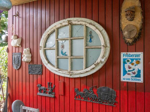An einer rot gestrichenen Holzwand hängen verschiedene Dekorationsgegenstände, darunter ein ovales Fenster mit antikem Rahmen, Schilder und Wandreliefs.