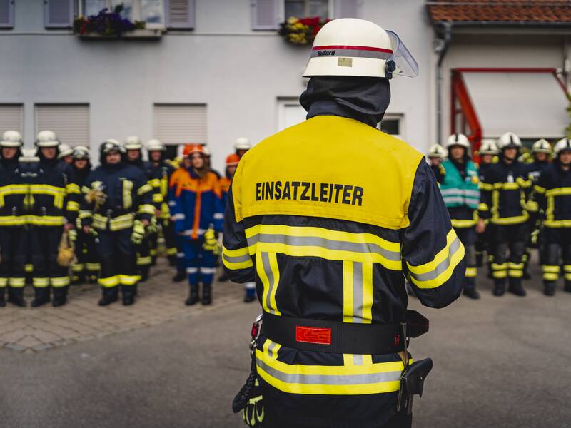 Im Vordergrund ist eine Person von hinten zu sehen mit einer Einsatzleiterjacke der Feuerwehr. Im Hintergrund sind mehrere Feuerwehrleute zu sehen.