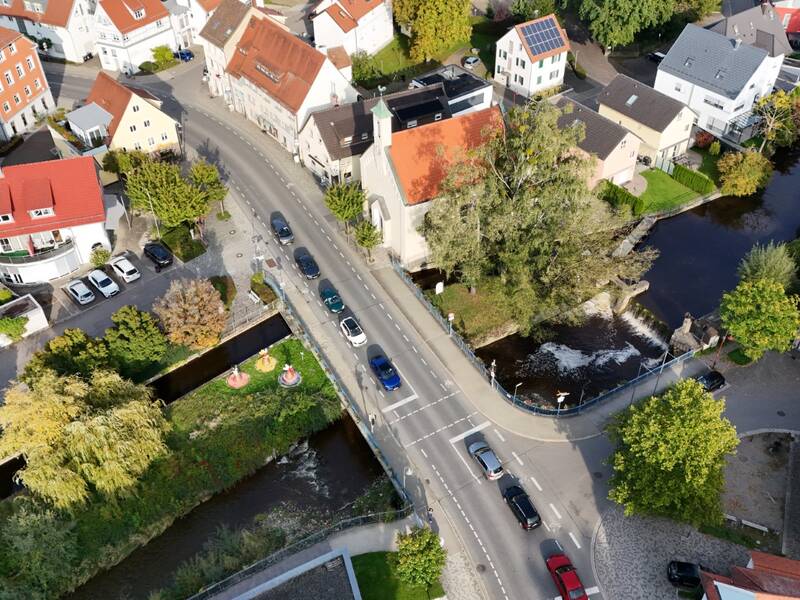 Eine Luftaufnahme zeigt eine Brücke mit Autos und Fußgängern, während daneben ein bewachsener Streifen mit bunten Skulpturen zu sehen ist.