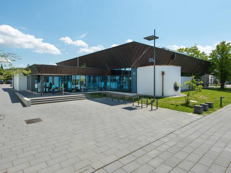 Das Bild zeigt ein modernes Gebäude mit großen Glasfenstern und einem markanten schrägen Dach, umgeben von grünen Rasenflächen und einem gepflasterten Vorplatz, auf dem einige Personen zu sehen sind.