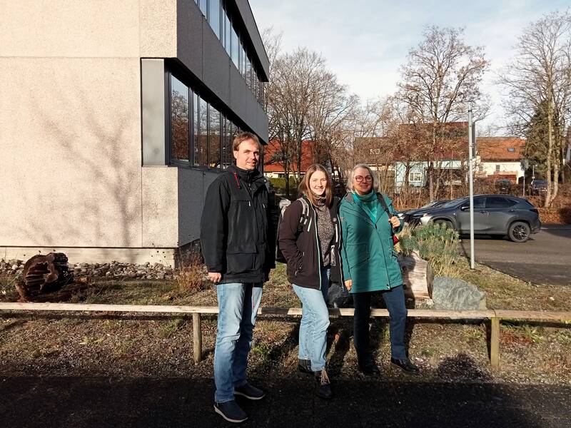 Drei Personen stehen draußen vor einem großen Gebäude, umgeben von kahlen Bäumen und einem geparkten Auto im Hintergrund.